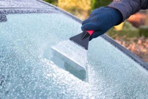 A gloved hand uses a small scraper to remove ice from a car windshield