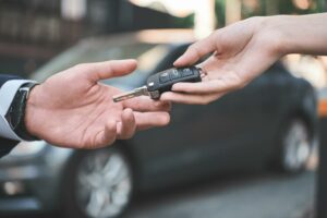 A person hands a car key off to another person, after selling their grey car in the background
