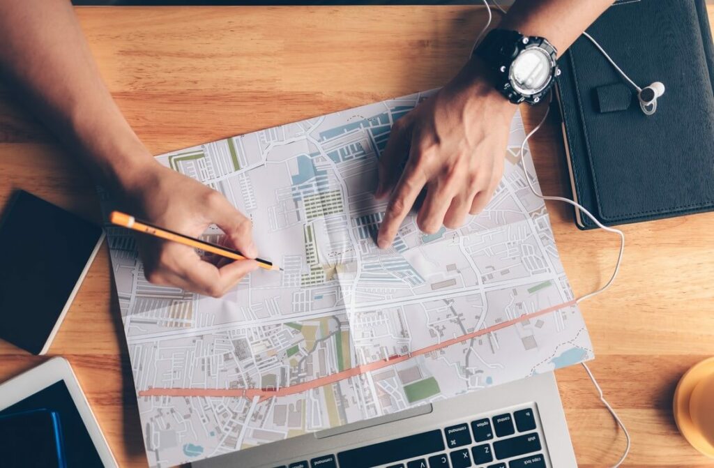 A person is drawing a route on the map on top of a desk with a laptop, notebook, and journal.