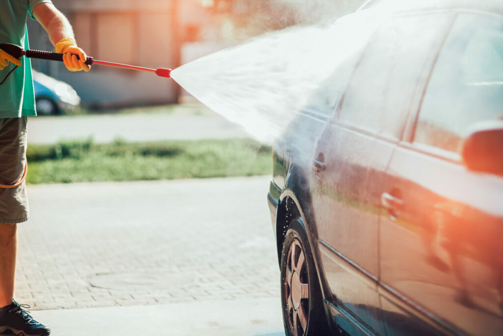 A man sprays his car with a hose as he washes it outside under the summer sun.
