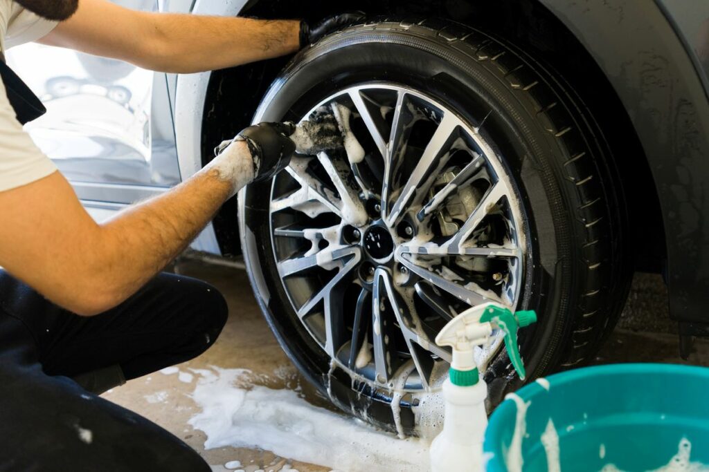 A close-up of a man washing a car tire using soap and a brush, with a spray bottle and a bucket beside him.