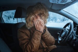 Man in down jacket tries to keep warm and not freeze in his stalled car in winter.