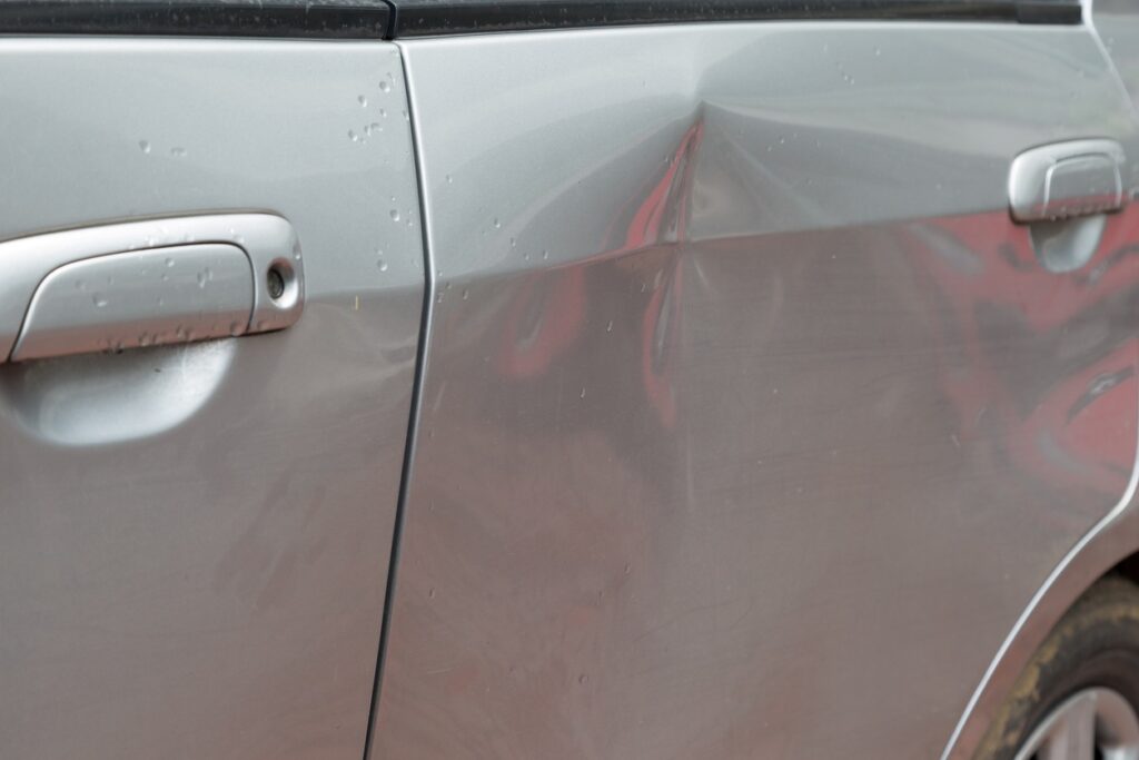 A silver car with a dent on the back door behind the driver’s seat.