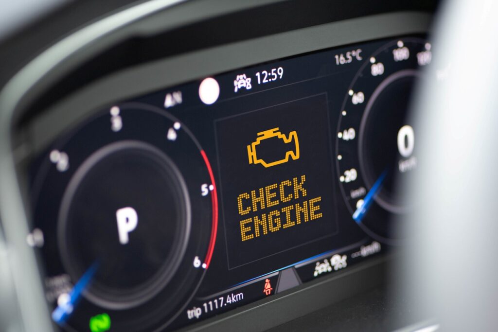 A digital dashboard in a modern car displaying an illuminated check engine light between the speedometer and odometer.
