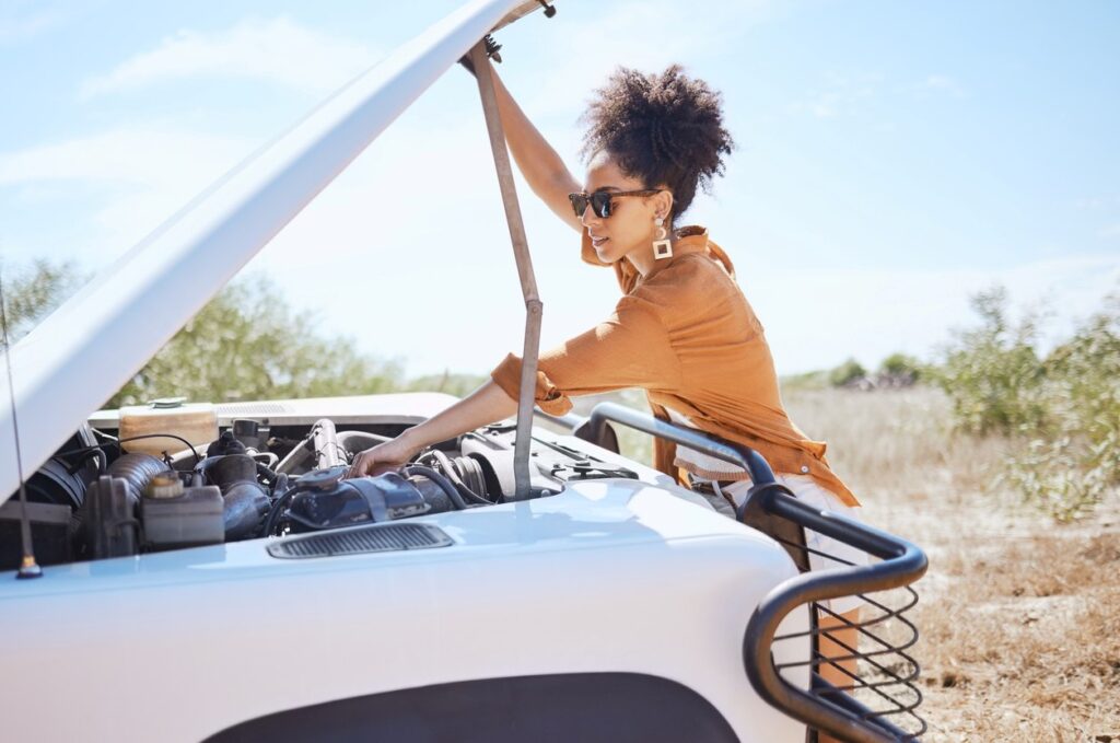 A young woman in sunglasses looks under the hood of a white car with a blue sky in the background.