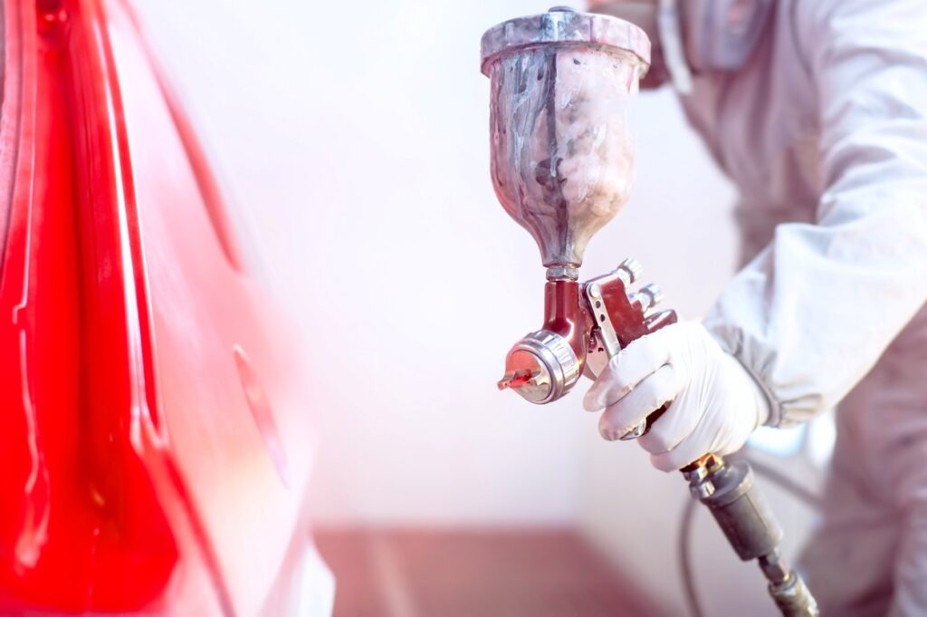 An auto body technician in a white jumpsuit and gloves applies a coat of red paint to a car using an airbrush.