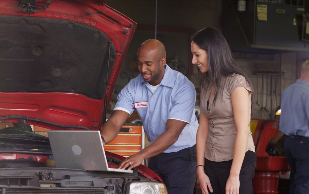 A car mechanic named Antonio in an auto shop works with a customer, showing his laptop to her.