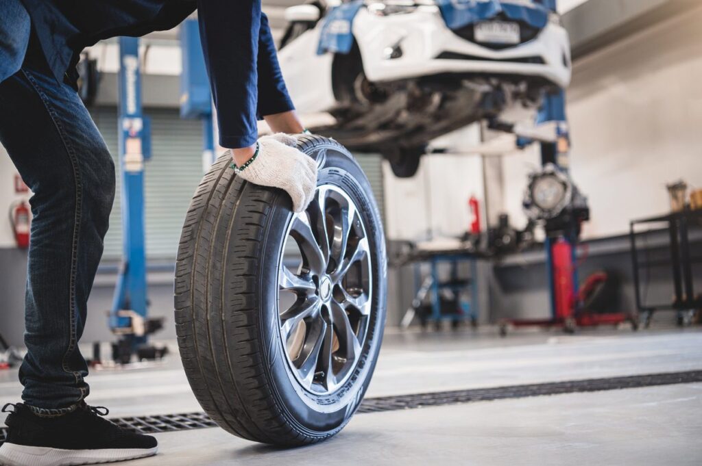 A mechanic in white gloves and a blue jumpsuit rolls a tire over to an elevated white car in an auto mechanic shop.