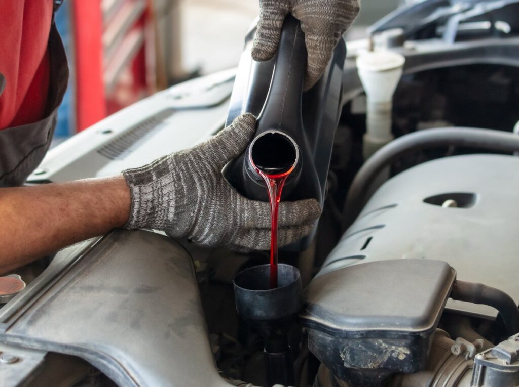 A mechanic in gloves holds a black plastic jug, pouring bright red liquid into a car component.