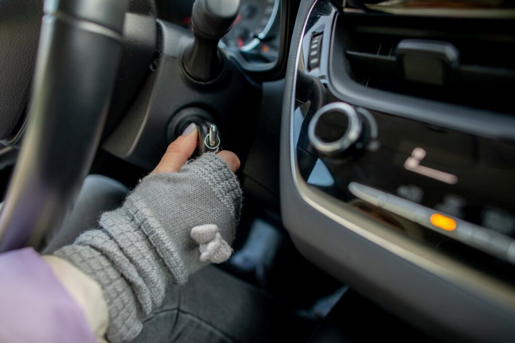 A hand in a gray glove, with gray nail polish, uses a car key to start the ignition behind the wheel of a car.