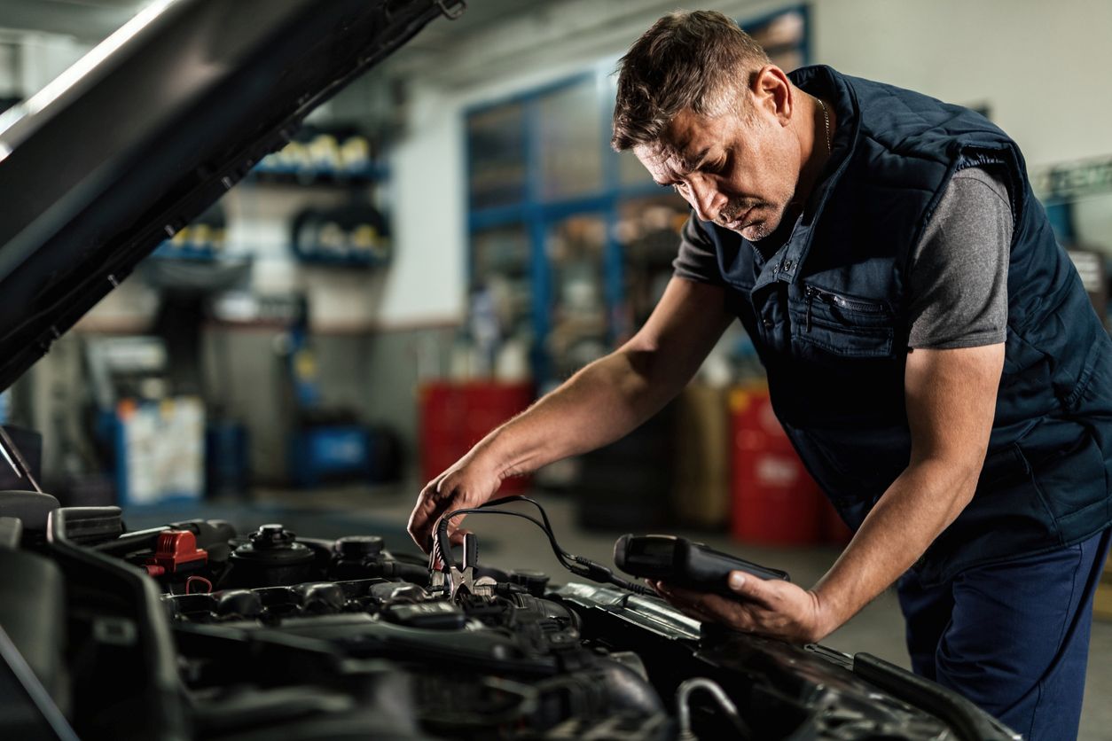 A mechanic testing a car battery with a handheld meter inside an auto repair shop.